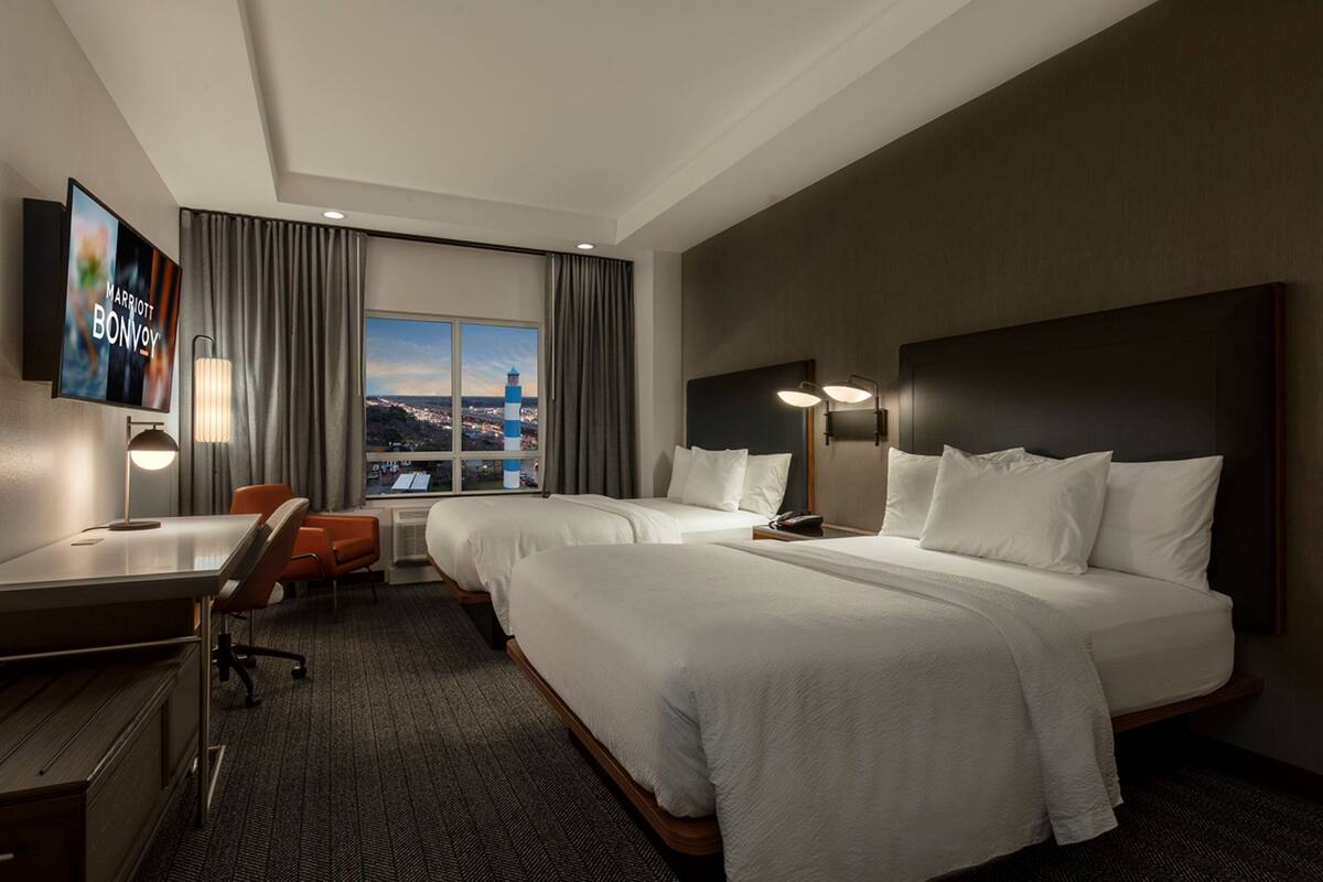 Ein Hotelzimmer mit zwei Betten, einem Schreibtisch, einem Sessel, einem Fernseher und Blick auf die Stadt.