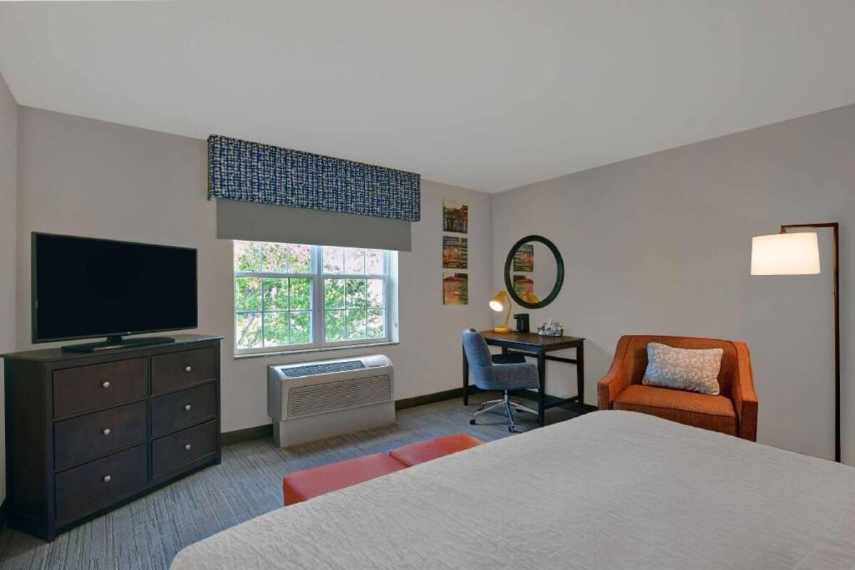 A hotel room with a bed, a desk, a chair, a television, a window, and a wall-mounted lamp.