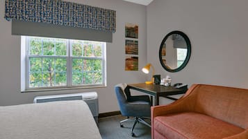 Una habitación de hotel con cama, escritorio y silla, sofá rojo, ventana con vistas a árboles y espejo en la pared.