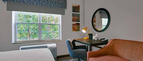A hotel room with a bed, a desk with a chair, a red sofa, a window with a view of trees, and a mirror on the wall.