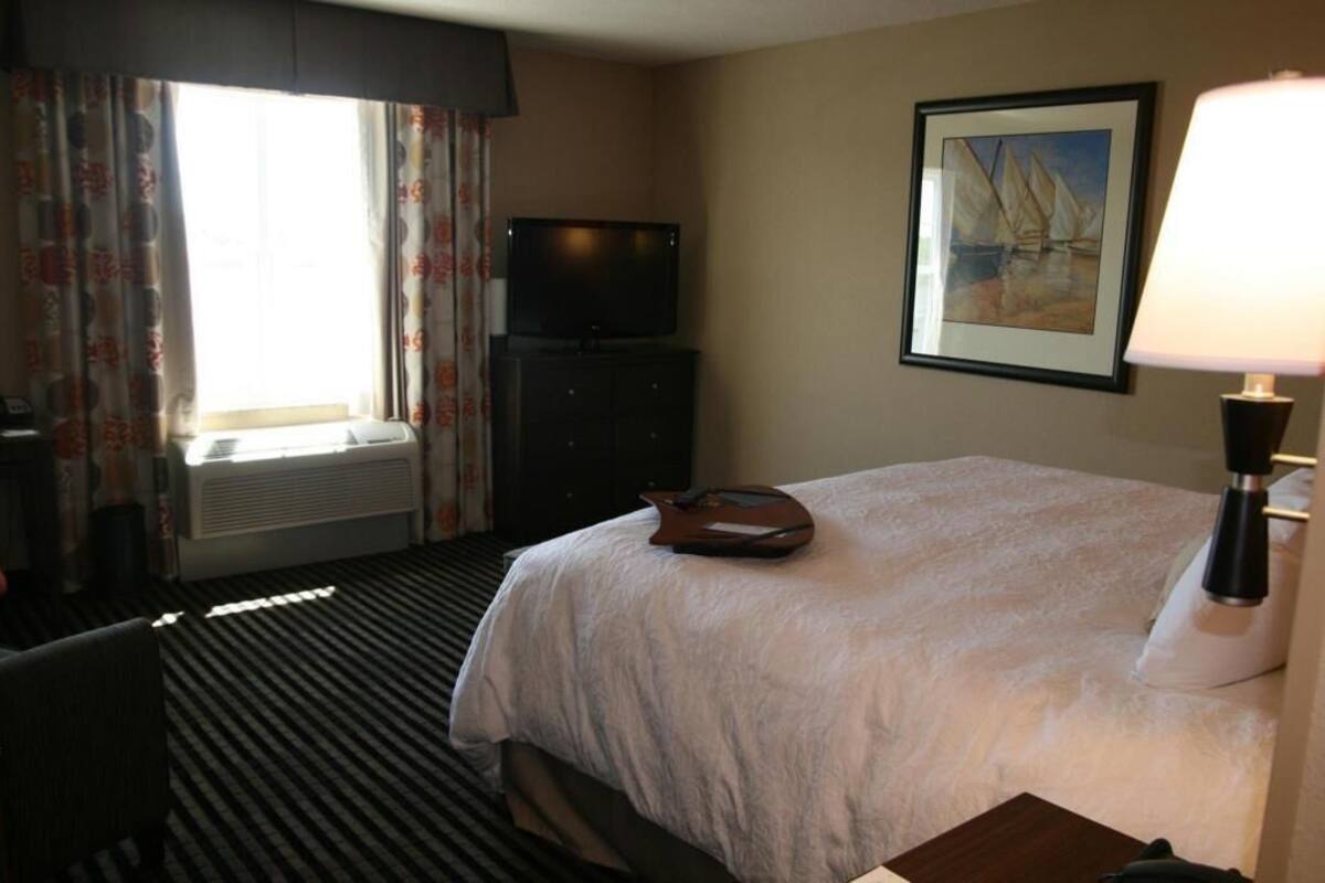 A hotel room with a bed, a television on a stand, a painting on the wall, and a lamp.
