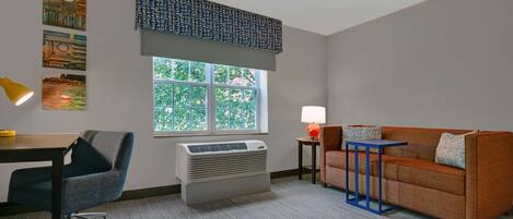 A room with a desk, chair, air conditioner, window, and a sofa with a lamp.