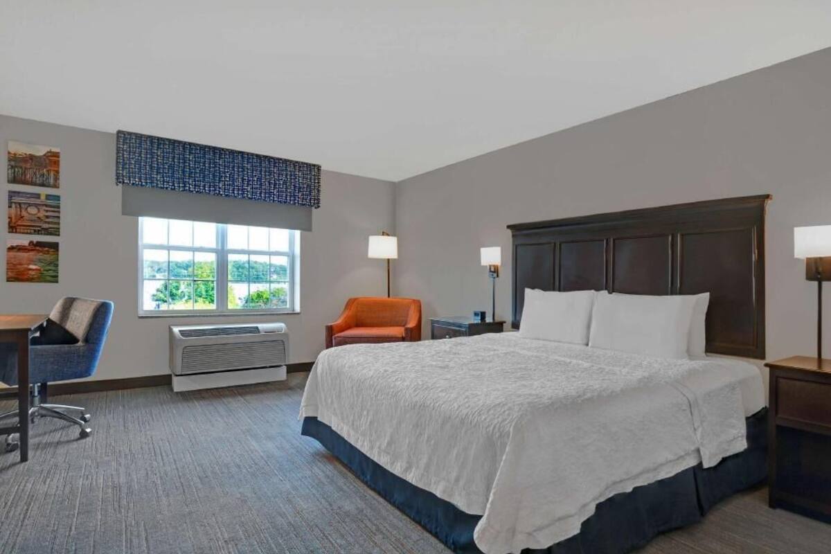 A hotel room with a large bed, a desk, an orange chair, a window with a view, and a wall-mounted air conditioner.
