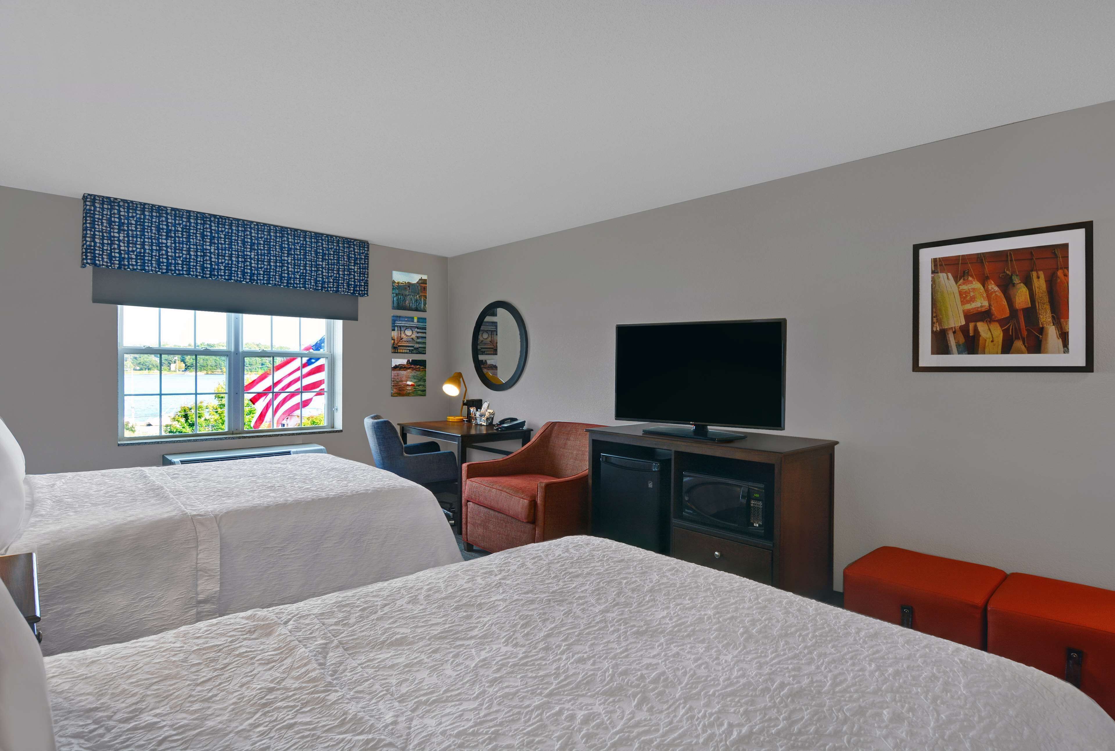 Et hotelværelse med to senge, et skrivebord med en computer, et fjernsyn og et vindue med et amerikansk flag.