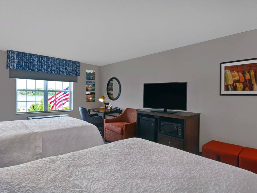 Ein Hotelzimmer mit zwei Betten, einem Schreibtisch mit Computer, einem Fernseher und einem Fenster mit amerikanischer Flagge.