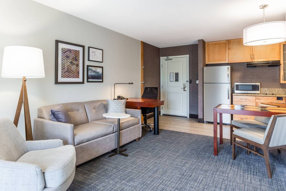 A modern living room with a sofa, a small round table, and a lamp. There is a kitchen area with a refrigerator, microwave, and wooden cabinets.