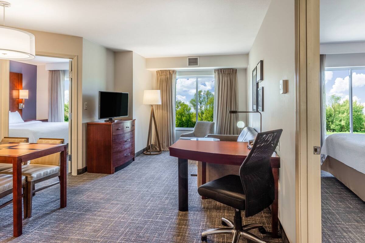 A hotel room with a bed, desk, chair, television, and a view of greenery.