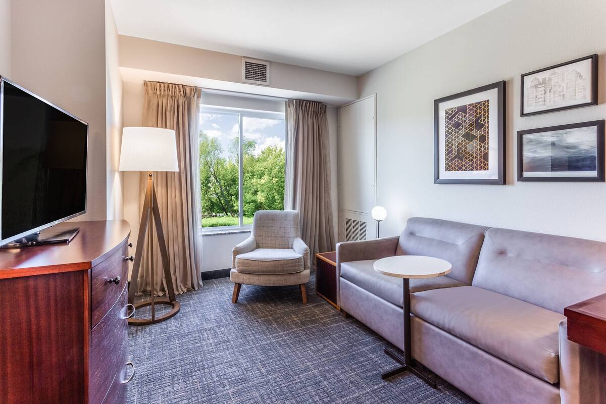 A hotel room with a flat-screen TV, a sofa, a chair, a lamp, and a window with curtains.