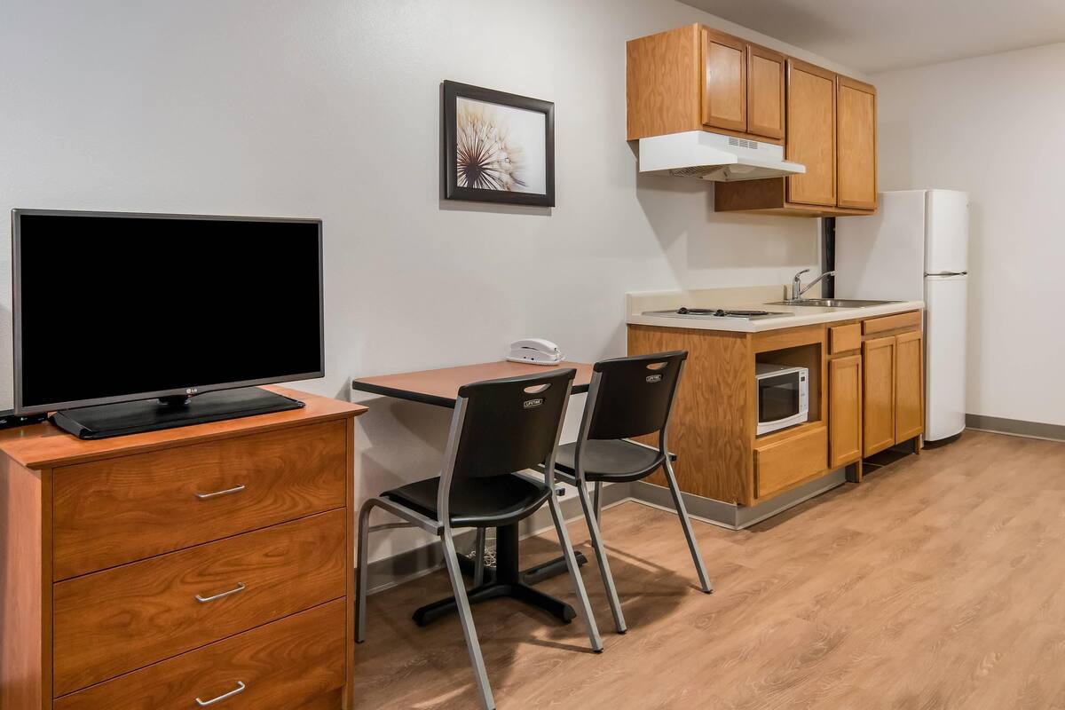 A kitchenette with wooden cabinets, a microwave, a refrigerator, a dining table with chairs, and a television on a wooden cabinet.