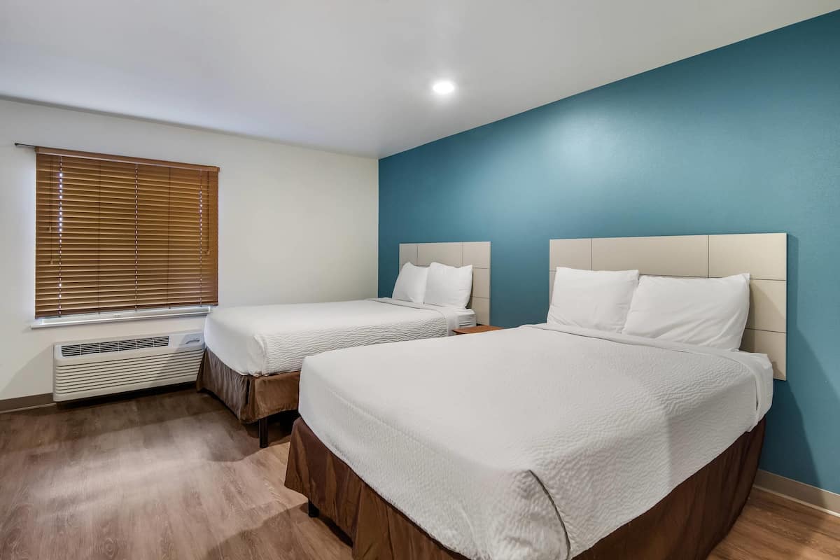 A room with two beds, a window with blinds, a wall heater, and a blue accent wall.