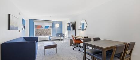 A modern hotel room with a large window, a blue sofa, a wooden dining table, and a desk with a chair.