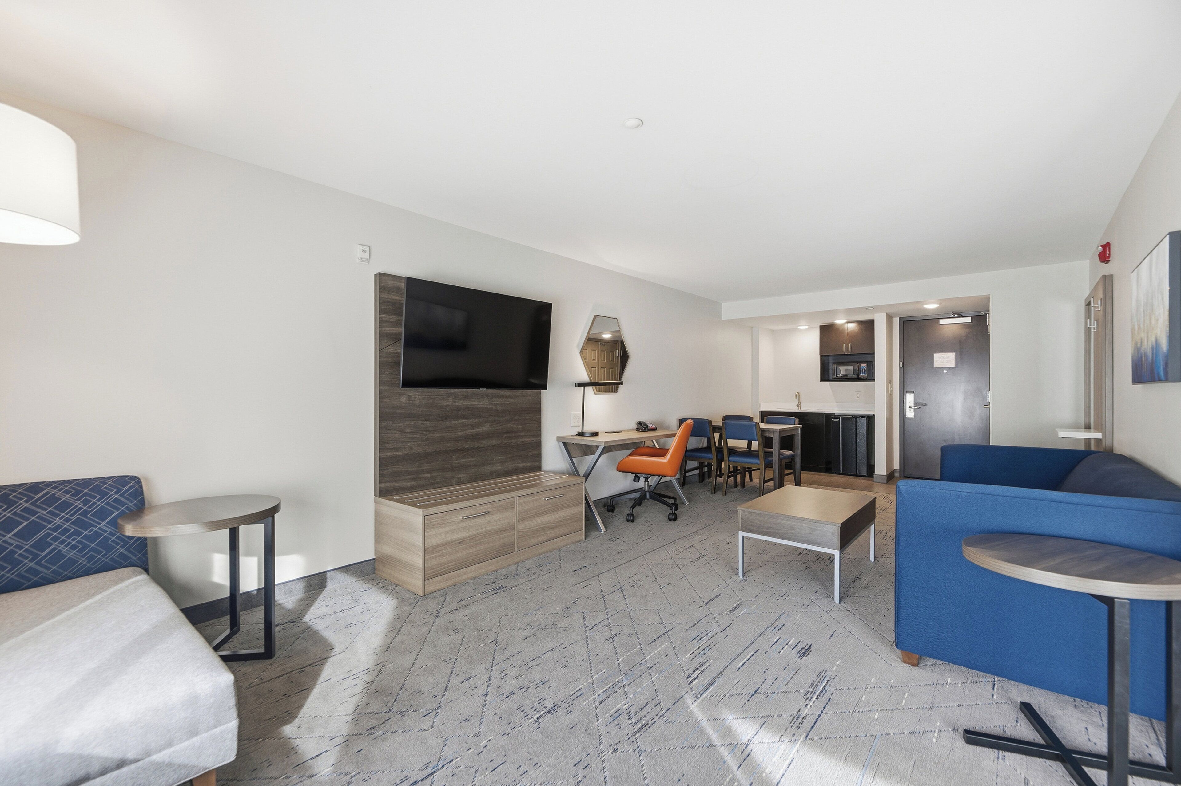 A modern hotel room with a sofa, a desk with a chair, a TV mounted on the wall, and a small table.