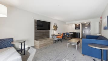 A modern hotel room with a sofa, a desk with a chair, a TV mounted on the wall, and a small table.