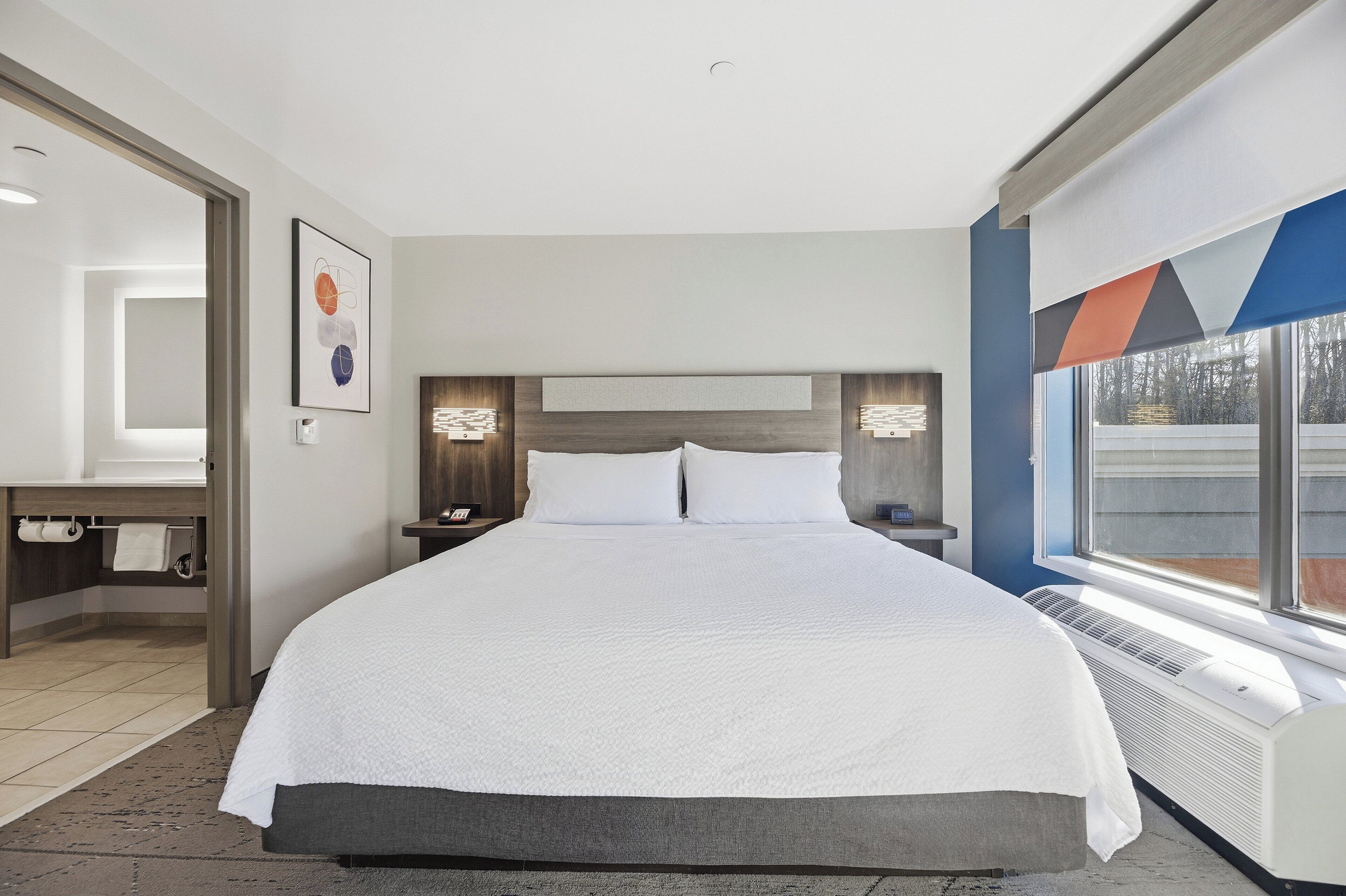 A modern hotel room with a large bed, a bedside table, a nightstand, a built-in shelf, a window with a view, and a bathroom with a mirror and towel rack.