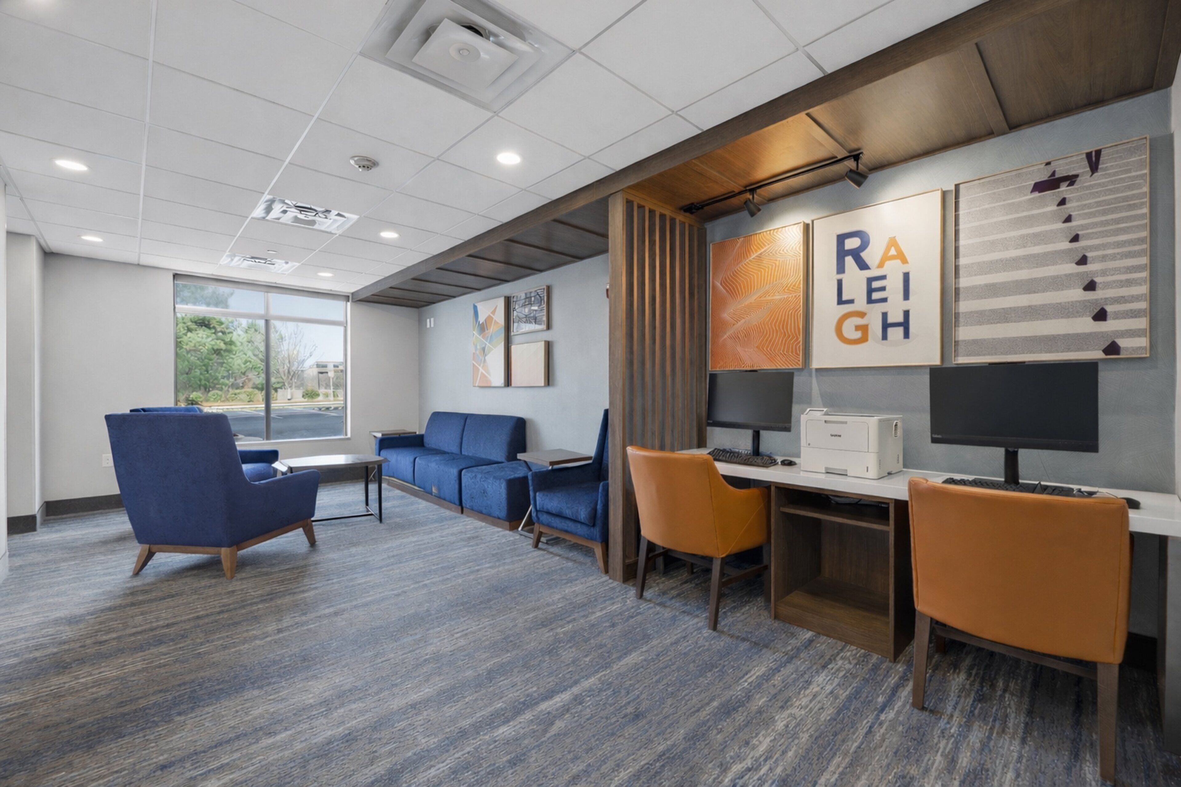 Un espace de bureau moderne avec un canapé bleu, des chaises orange, un bureau avec un ordinateur et des œuvres d’art encadrées accrochées au mur.