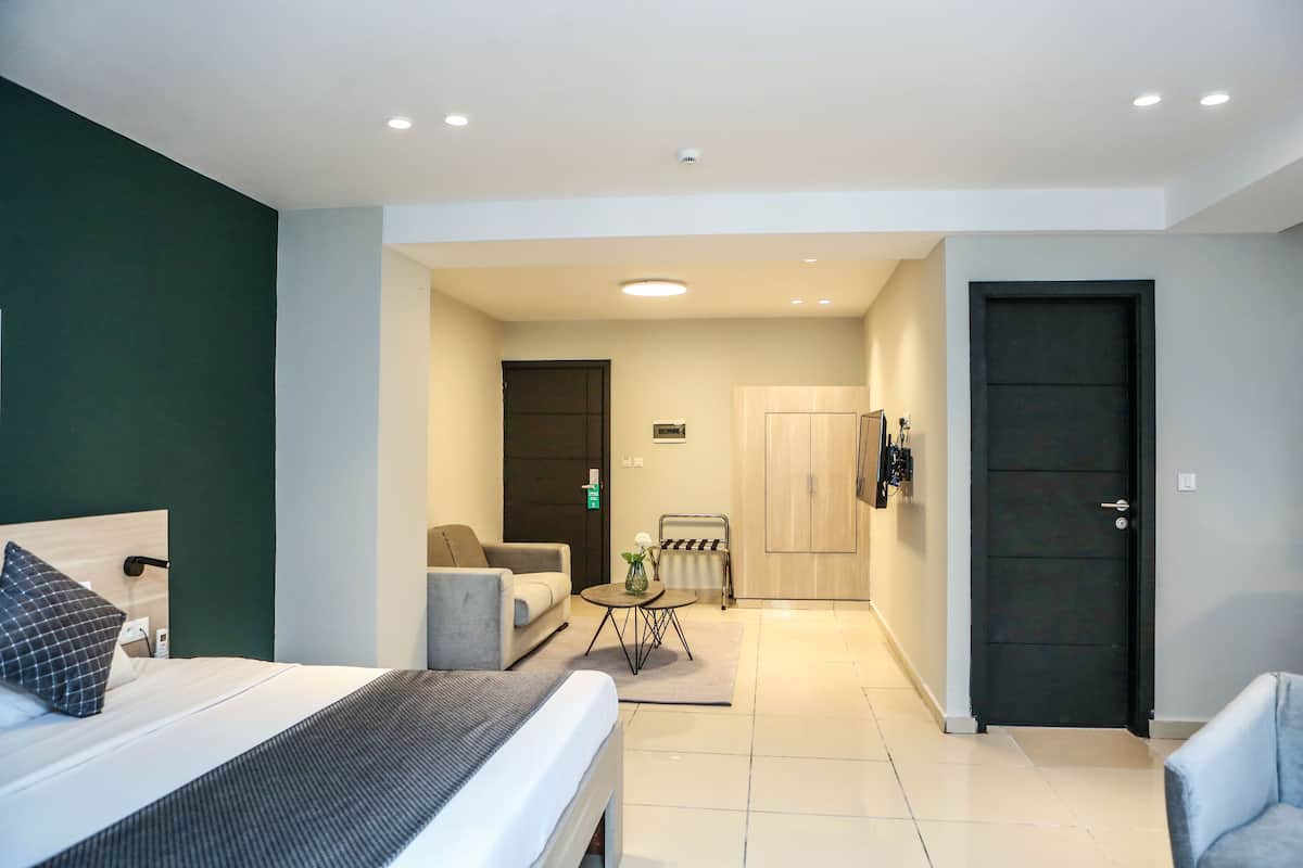 A modern hotel room with a bed, a sofa, a small table with a vase, and a door leading to a bathroom.