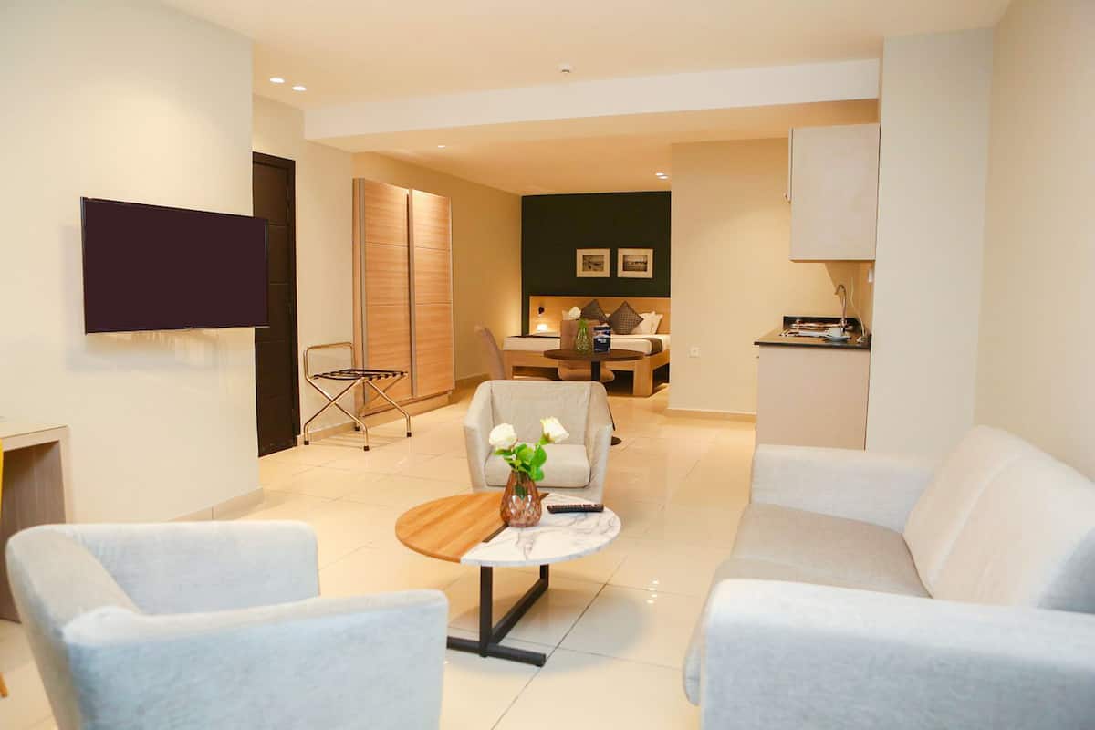 A modern hotel room with a flat-screen TV, a dining area with a table and chairs, a sofa, and a small coffee table with a vase of flowers.