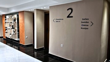 Un pasillo moderno de hotel con un cartel con el número '2', que indica el segundo piso. Flechas direccionales señalan 'Chambres 208-211' y 'Salles Samba Ebène Kosipo'.