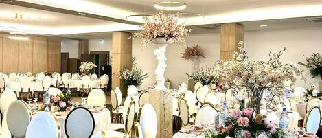 Un salón de banquetes con mesas redondas, sillas blancas y centros de mesa florales.