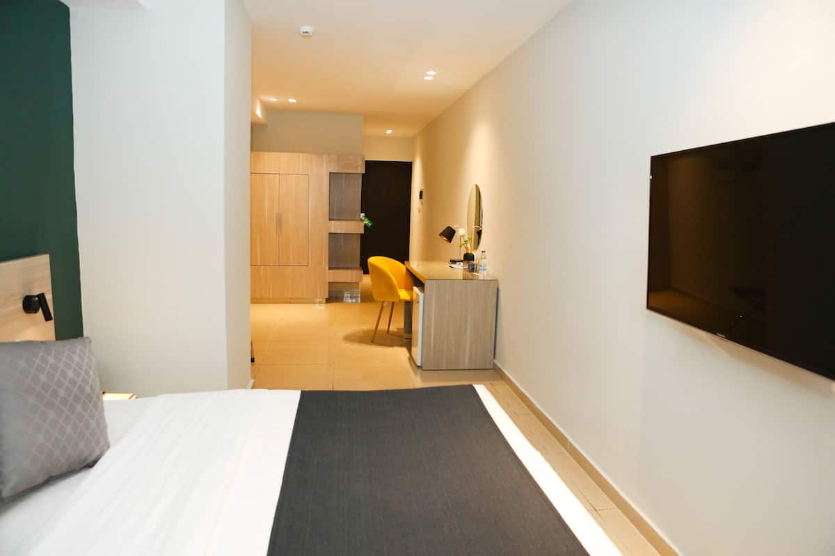 A hotel room with a bed, a desk with a chair, a TV, and a small kitchenette in the background.