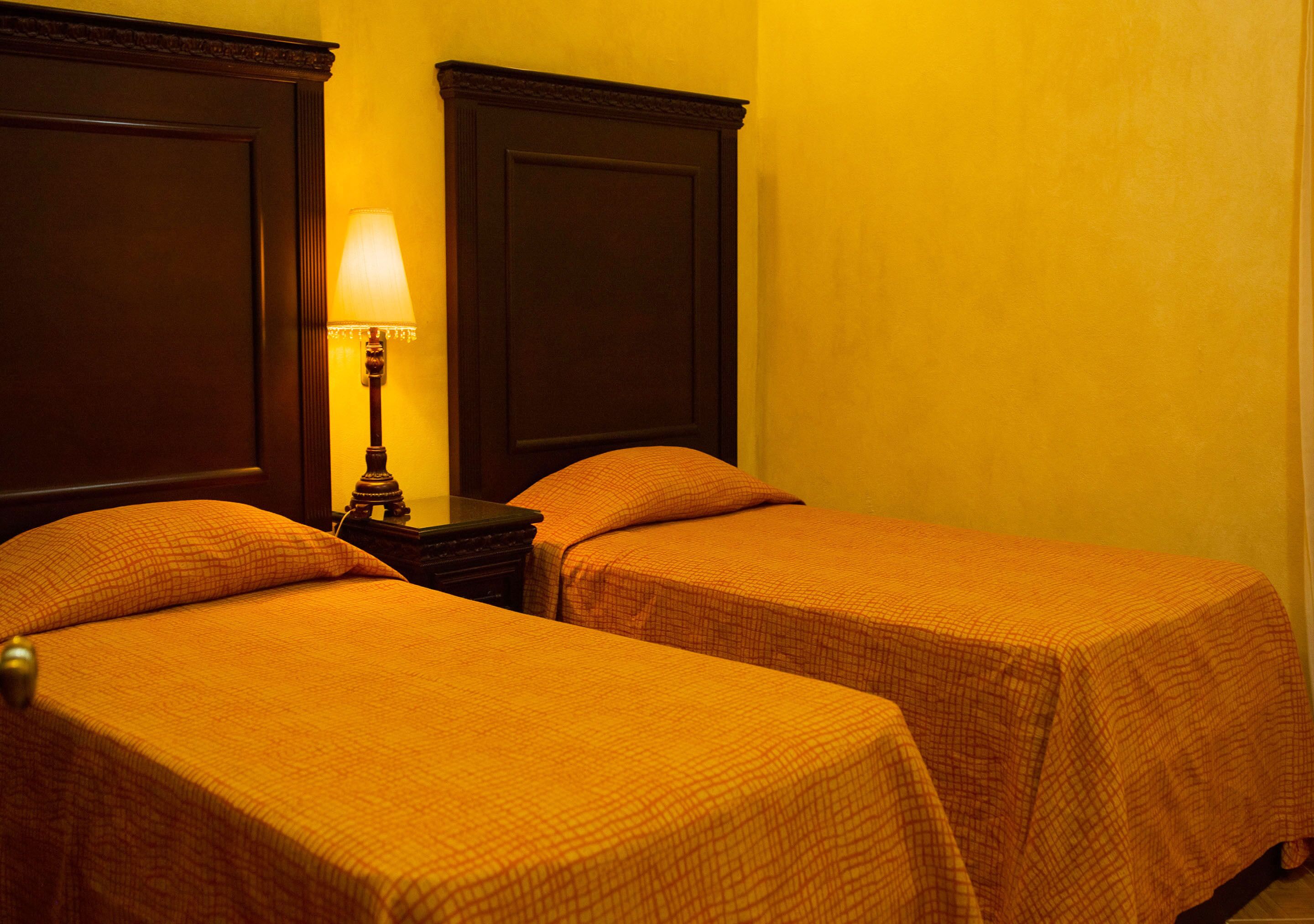 A hotel room with two beds, each with a wooden headboard and a checkered bedspread. A nightstand with a lamp is between the beds.