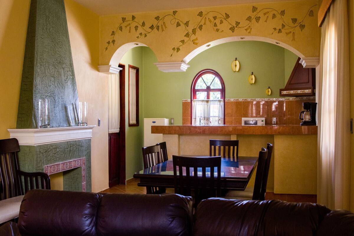 A dining area with a table and chairs, a fireplace, and a kitchen area with a window and a hanging light fixture.