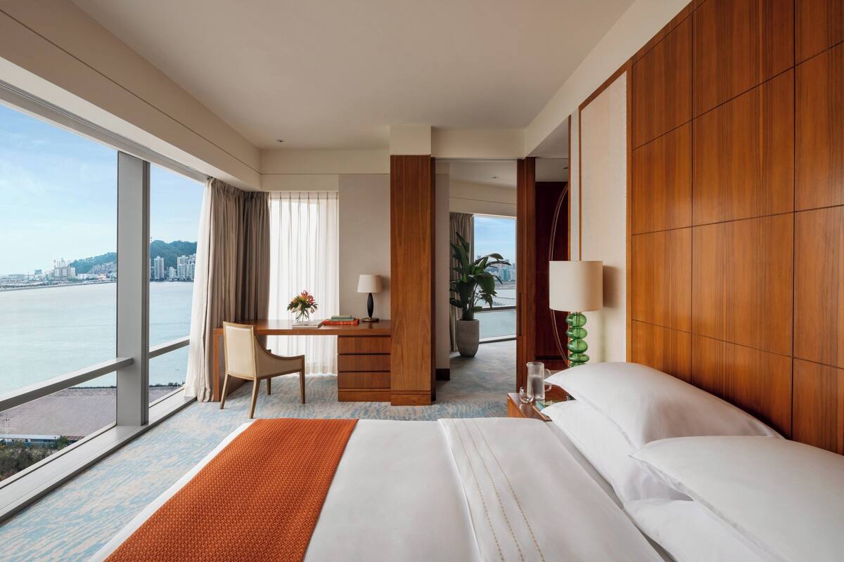 A hotel room with a large bed, a desk with a chair, a view of the water, and a cityscape.