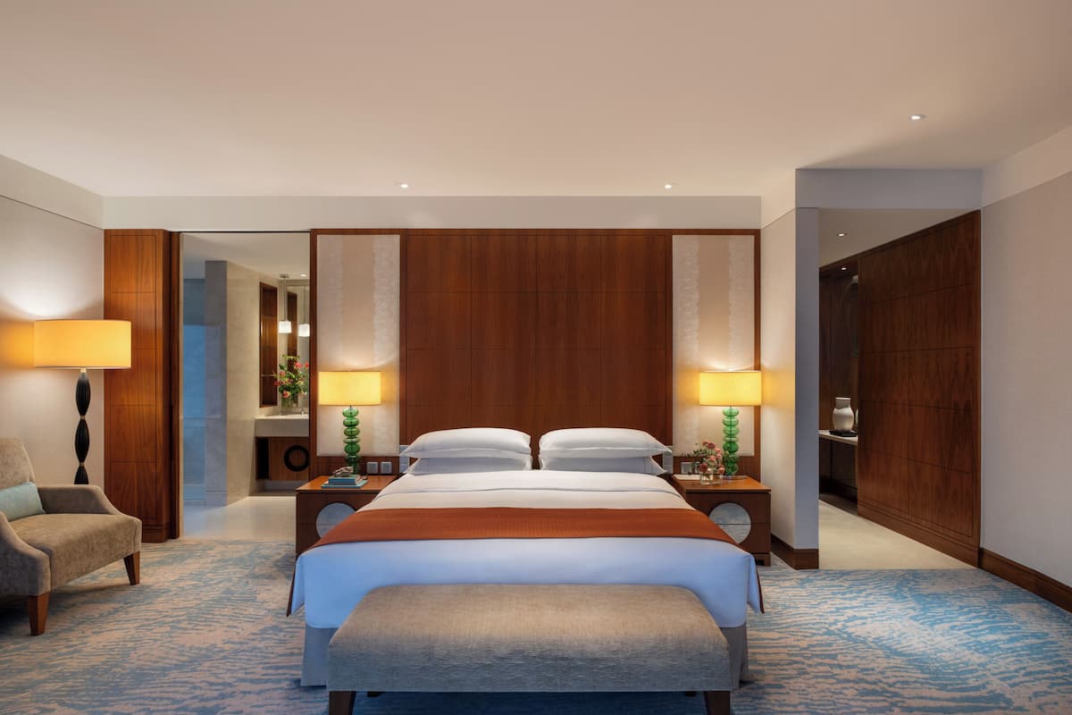 A hotel room with a large bed, two bedside tables with lamps, a bench, a chair, and a mirror.