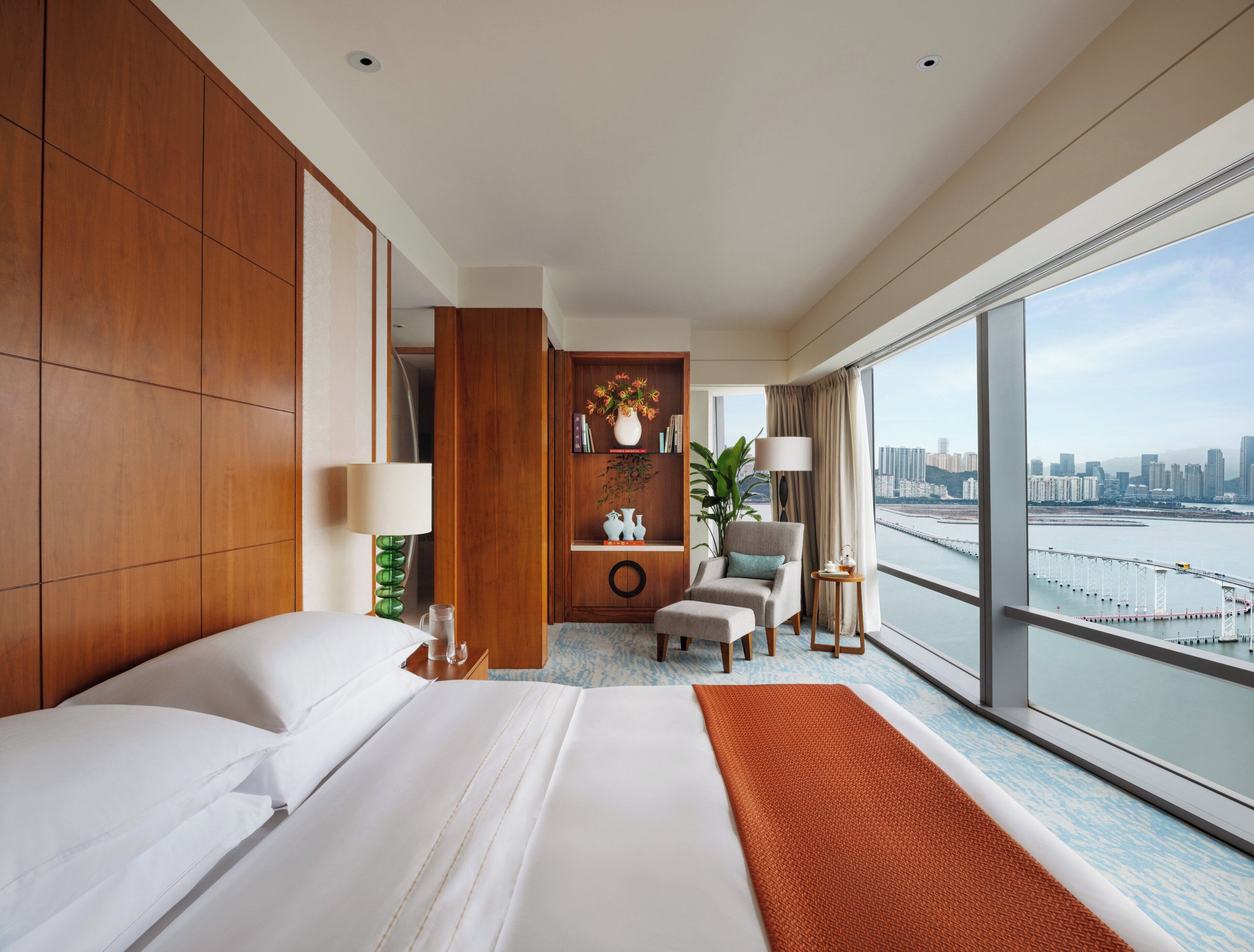 Habitación de hotel con una cama grande, un área de descanso con una silla y una mesa, una estantería y vistas a un horizonte urbano a través de amplias ventanas.