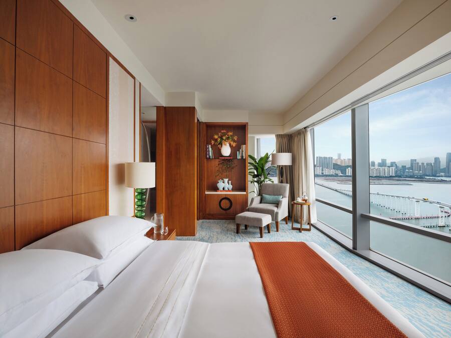 Habitación de hotel con una cama grande, un área de descanso con una silla y una mesa, una estantería y vistas a un horizonte urbano a través de amplias ventanas.