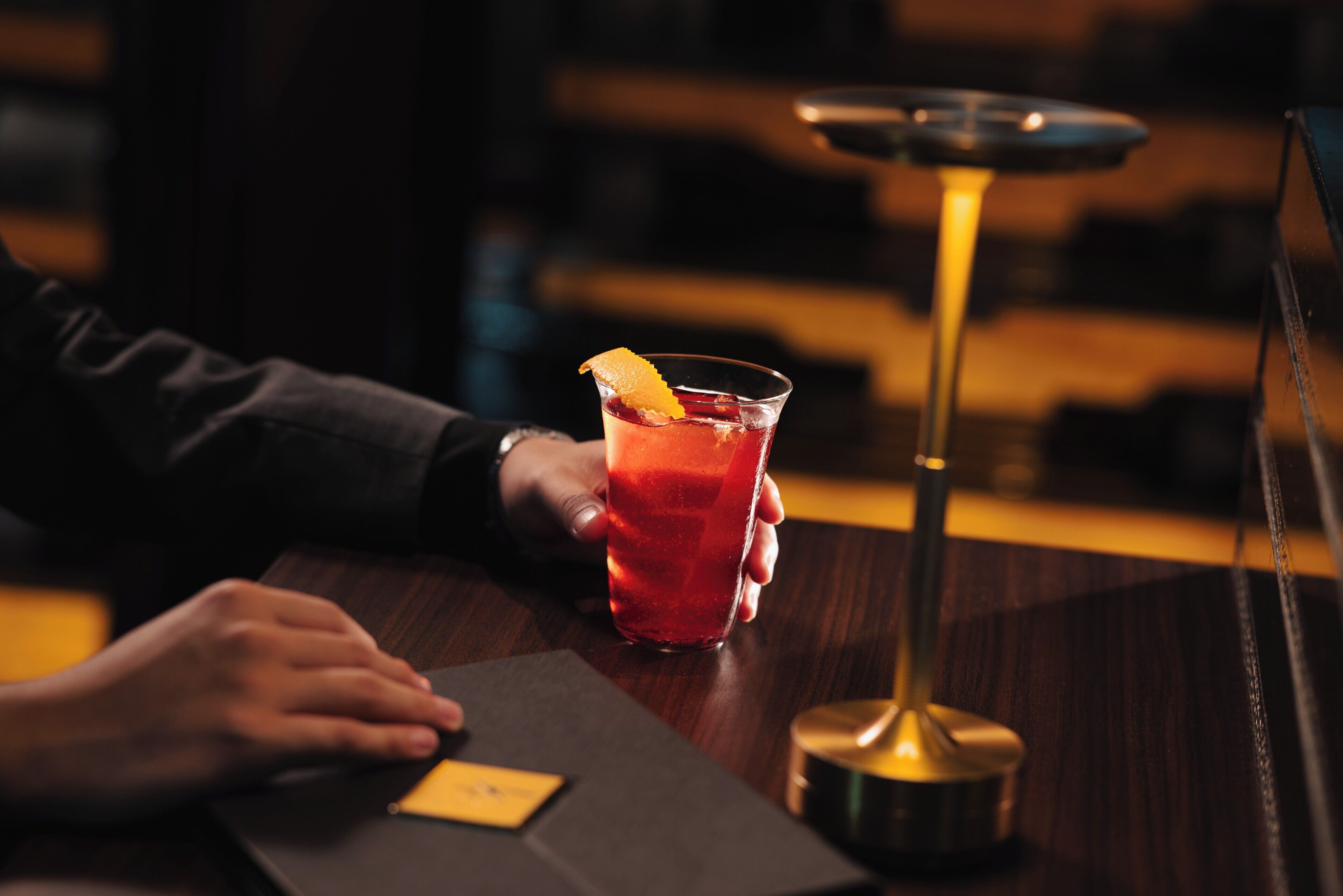 A person holding a glass with a garnish, a bar stool, and a bar counter.