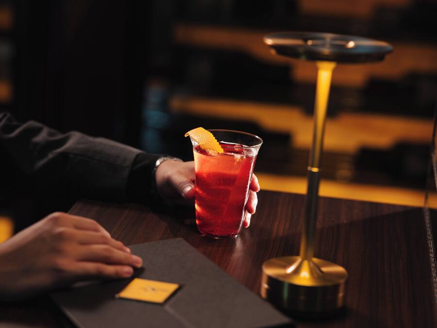 A person holding a glass with a garnish, a bar stool, and a bar counter.