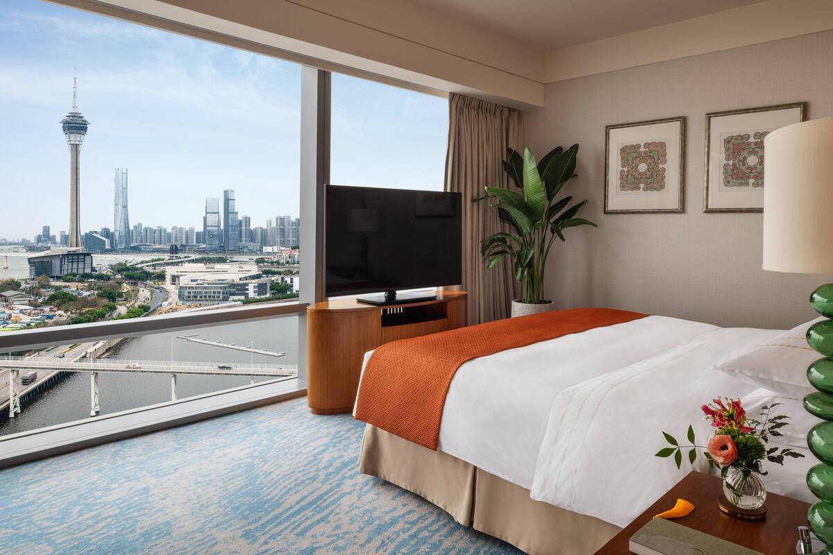 A hotel room with a large window, a bed, a TV, a plant, and artwork on the wall.