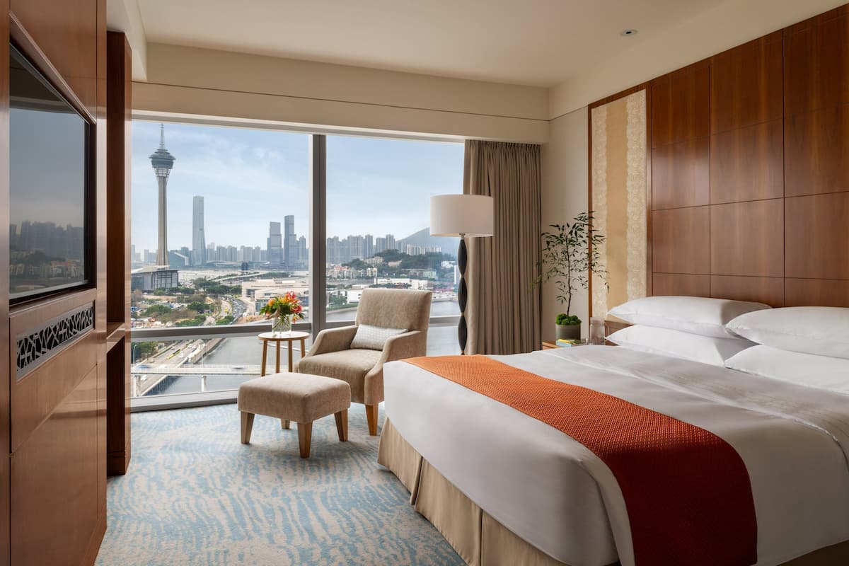 A hotel room with a large bed, a TV, a desk, and a view of a cityscape including a tower.