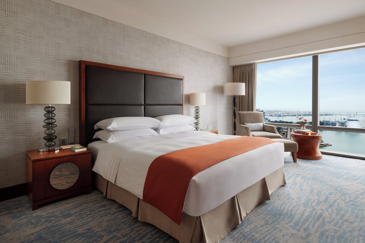 A hotel room with a large bed, two bedside tables, a chair, and a view of a cityscape through a large window.