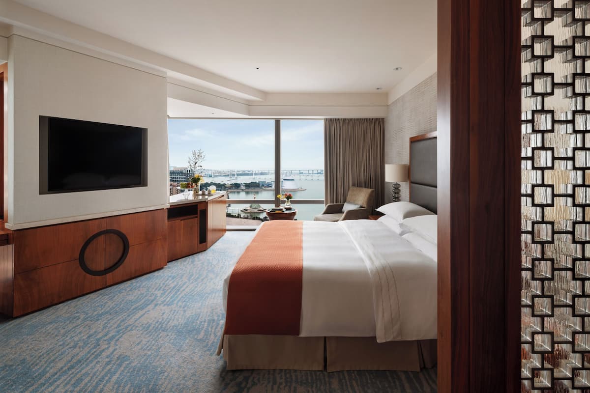 A hotel room with a large bed, a flat-screen TV, a desk, and a view of a bridge and water.