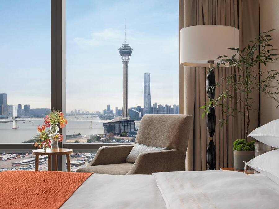 A hotel room with a bed, a chair, a lamp, and a view of a cityscape including a tower.