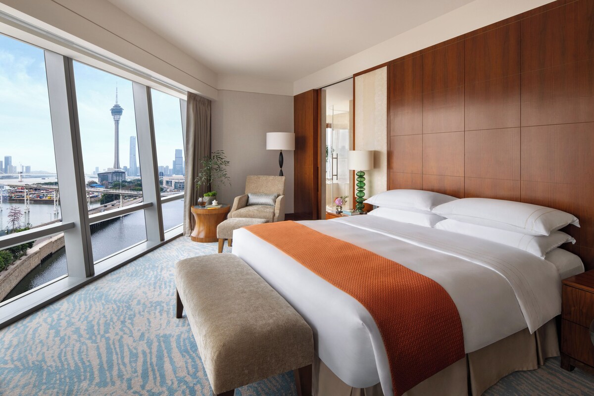 A hotel room with a large bed, a bench, a chair, a desk, and a view of a cityscape.