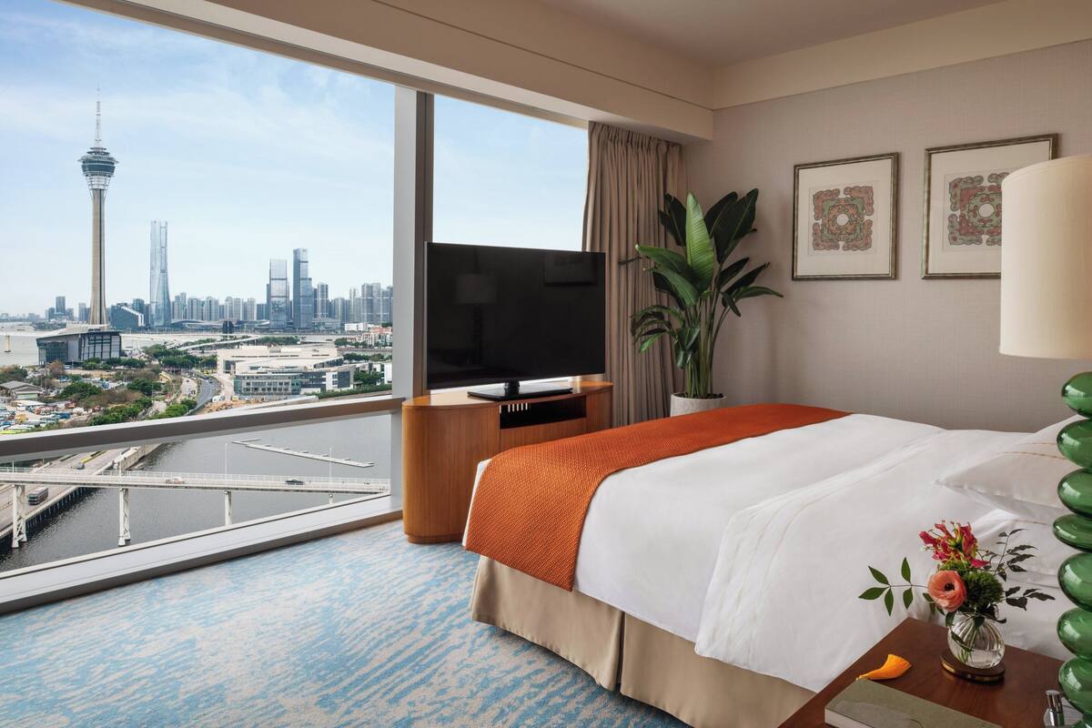 A hotel room with a large window, a bed, a TV, a plant, and artwork on the wall.