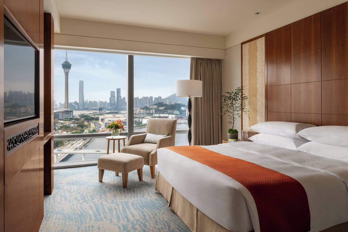 A hotel room with a large bed, a TV, a desk, and a view of a cityscape including a tower.