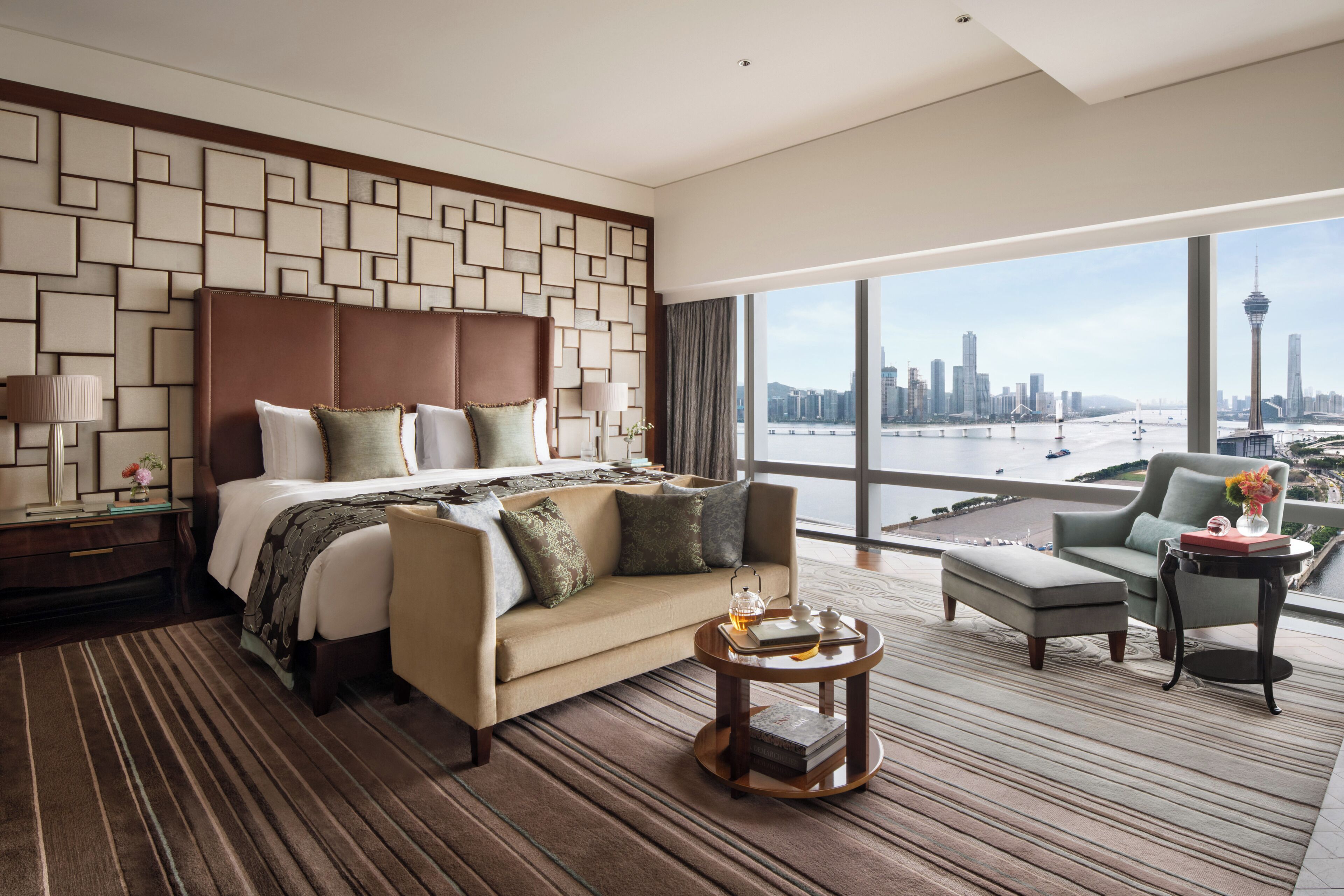 Habitación de hotel con una cama grande, un sofá, una mesita y vistas a un horizonte urbano.