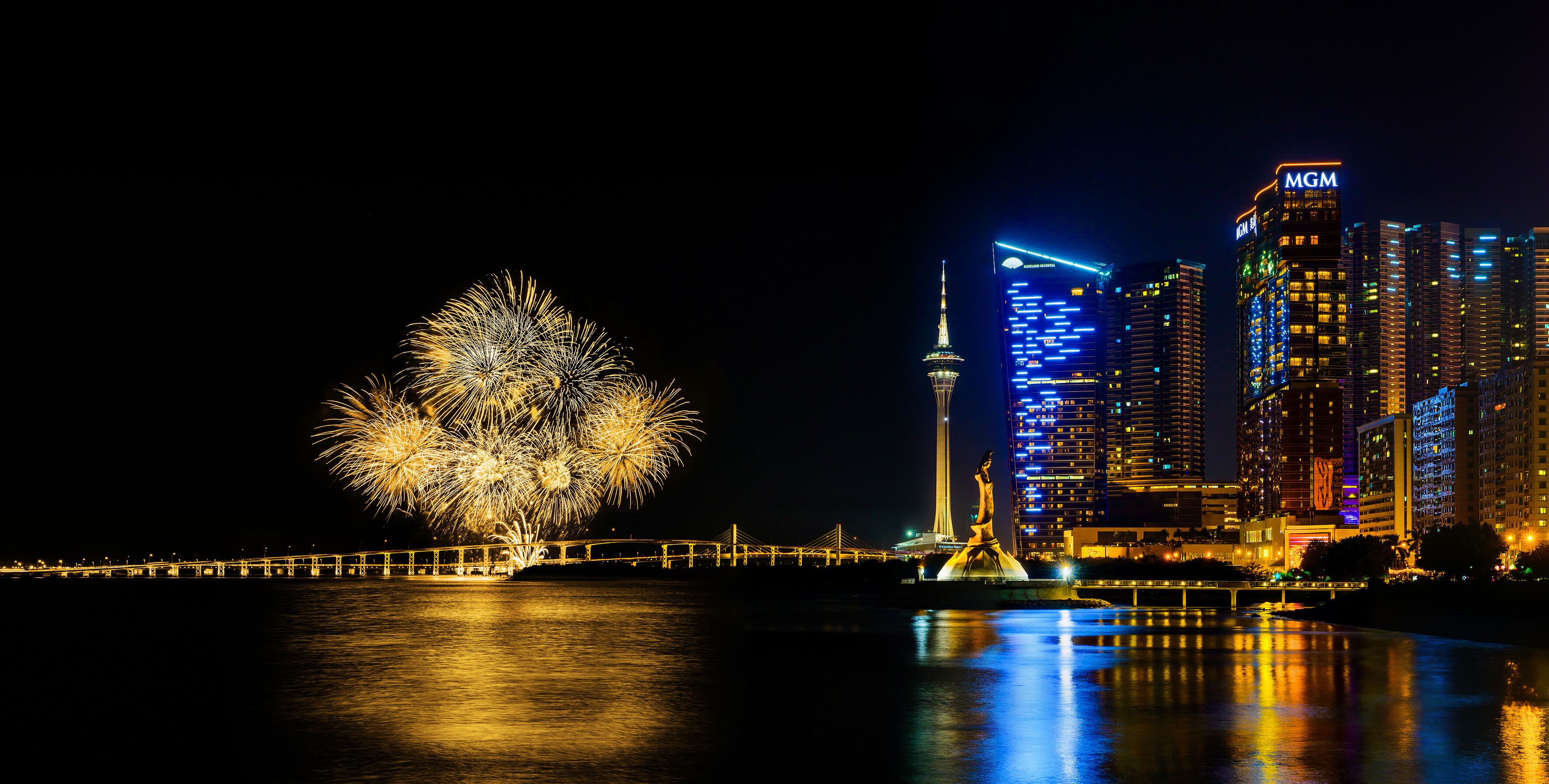 A nighttime cityscape with illuminated buildings, a prominent tower, and fireworks over a body of water.