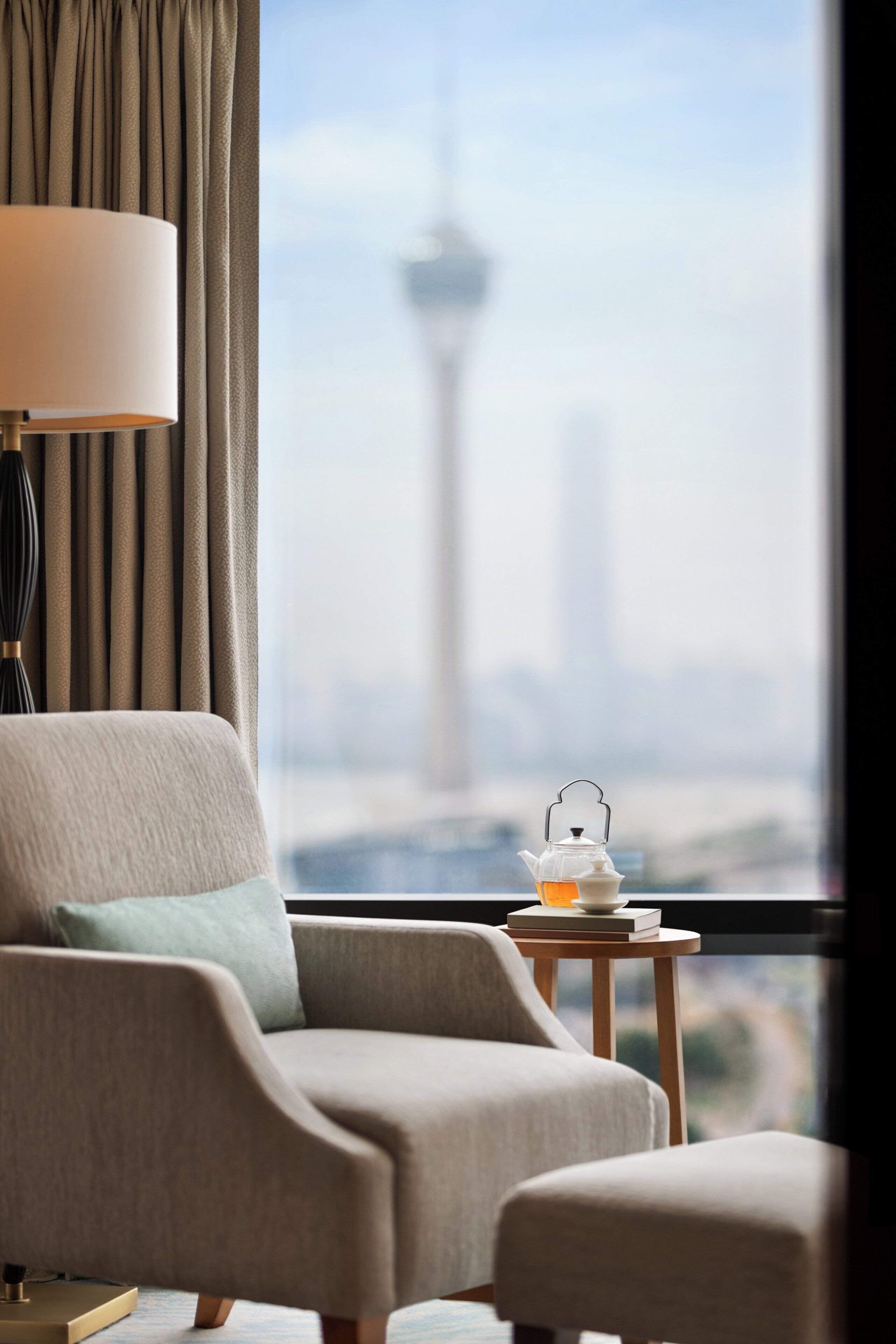 A hotel room with a view of a tower, a chair, a table with a teapot, and a lamp.
