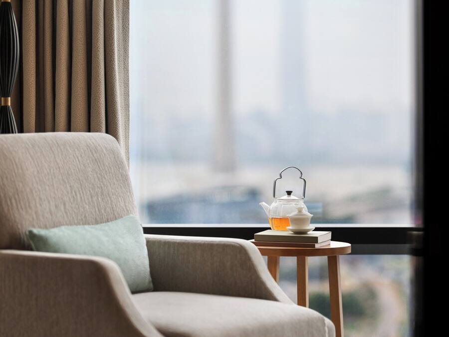 A hotel room with a view of a tower, a chair, a table with a teapot, and a lamp.