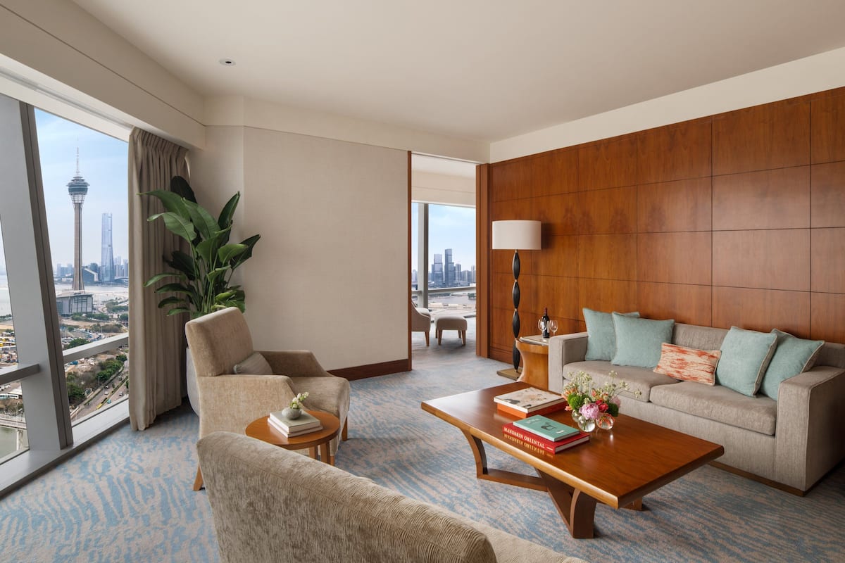A hotel room with a large window offering a city view, a sofa, a coffee table, and a side table with books and a vase of flowers.