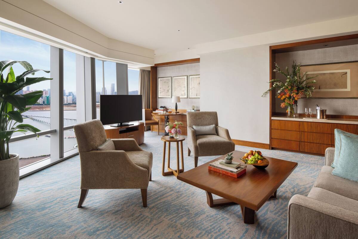 A spacious living room with large windows, a flat-screen TV, comfortable seating, a coffee table, and a view of the city skyline.