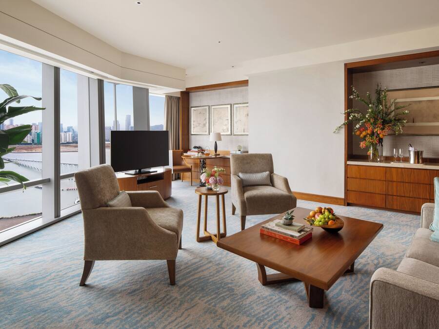 Una habitación de hotel moderna con un ventanal grande, un televisor de pantalla plana, dos sillones, un sofá, una mesa de centro y una zona de comedor con una mesa pequeña y sillas.