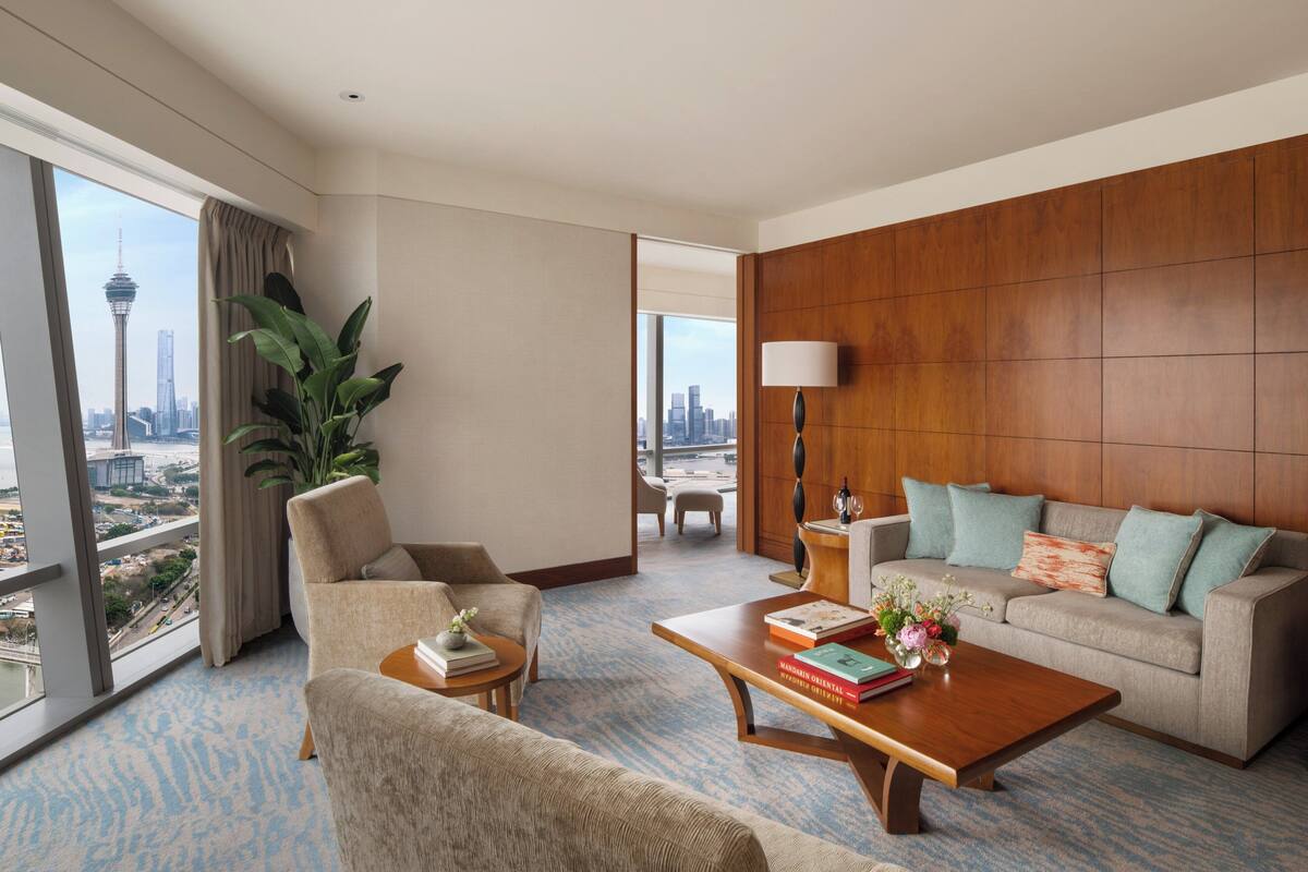 A hotel room with a large window offering a city view, a sofa, a coffee table, and a side table with books and a vase of flowers.