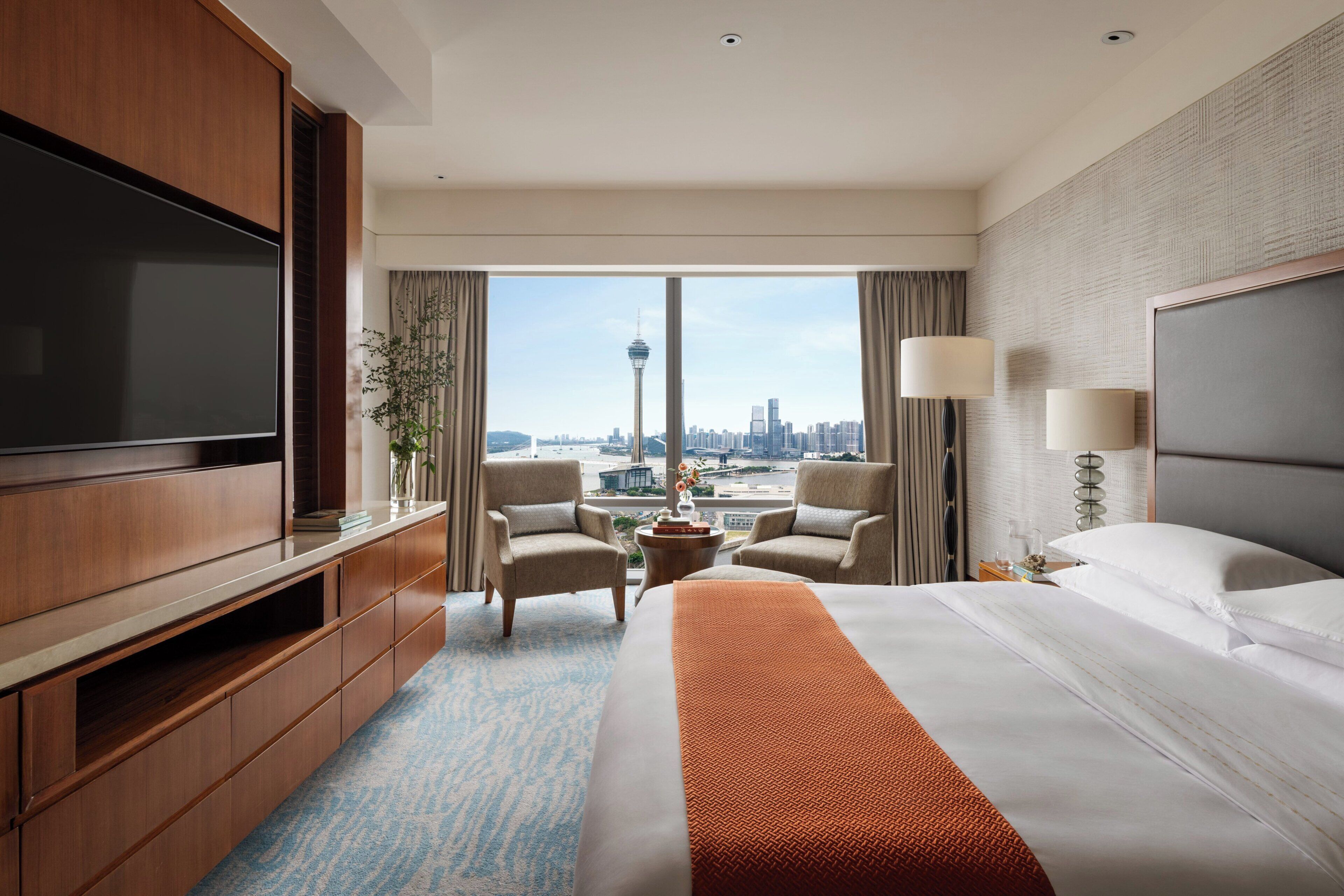 A hotel room with a bed, a TV, a desk, and a view of a cityscape.