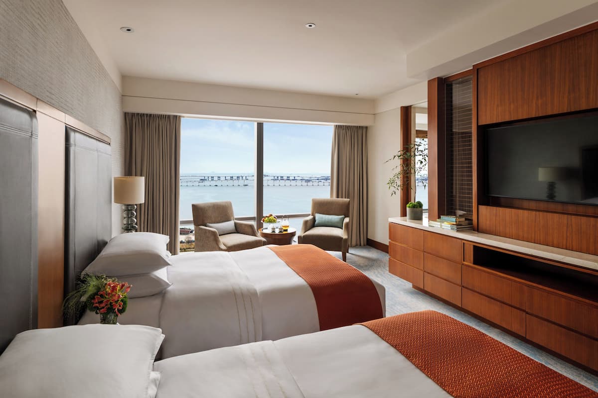 A hotel room with two beds, a TV, a desk with a lamp, and a view of the sea.